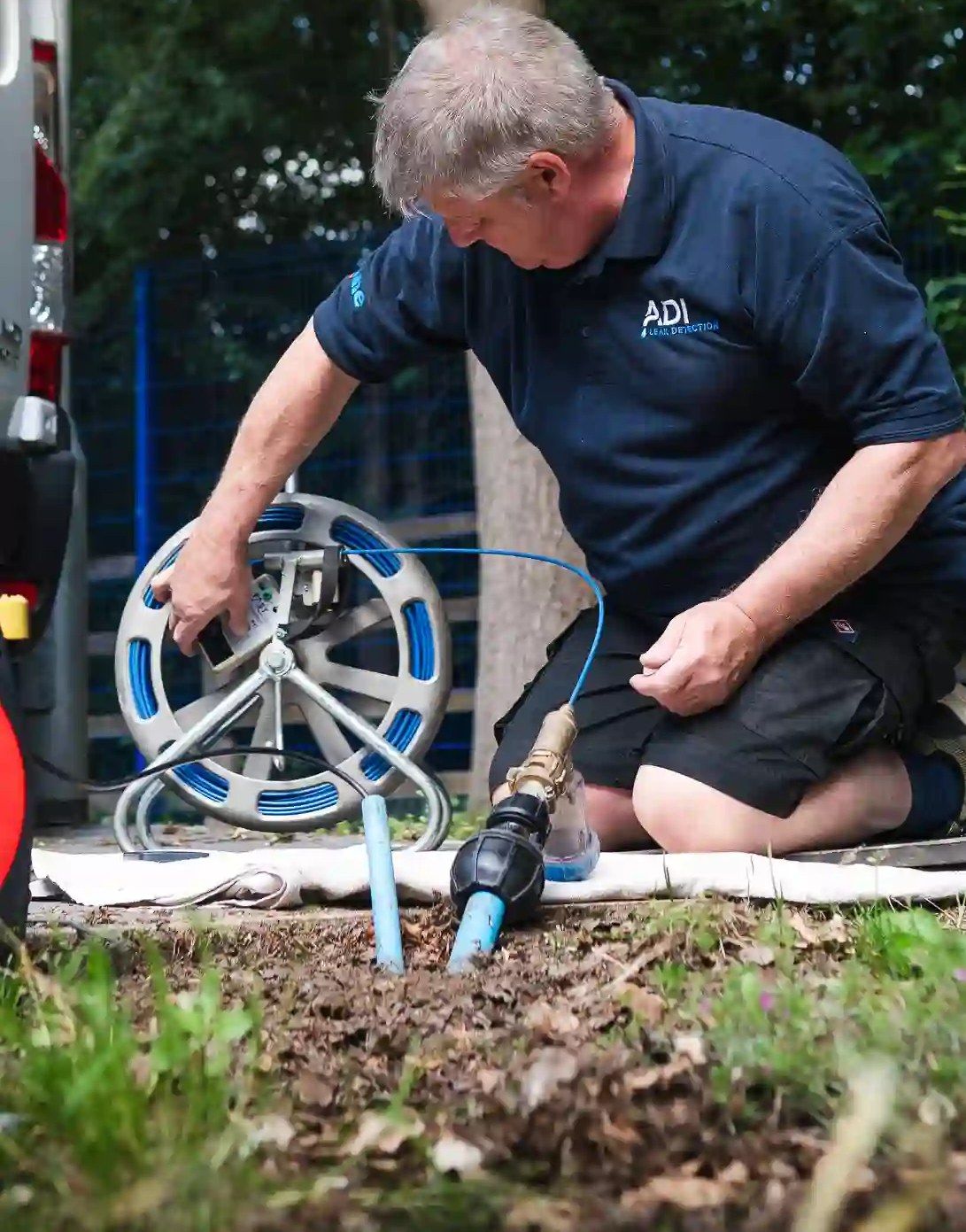 An engineer kneeling in a front garden, connecting test equipment to a blue water supply pipe
